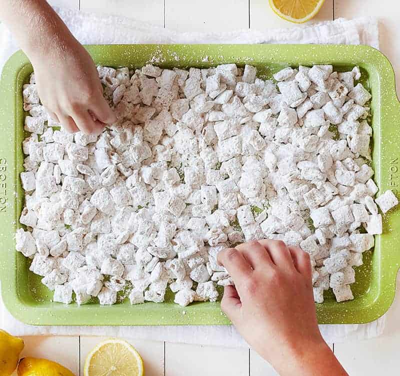 Lemon Puppy Chow (aka Muddie Buddies) is a super fun citrus twist on a classic! Sweet vanilla and lemon flavor all wrapped up in an easy-to-eat snack! #lemonpuppychow #lemonmuddiebuddies #puppychow #puppychowrecipe #muddiebuddies #easysnacks #snacks #afterschool #iambaker #nobake 孩子们的手正在吃柠檬狗粮