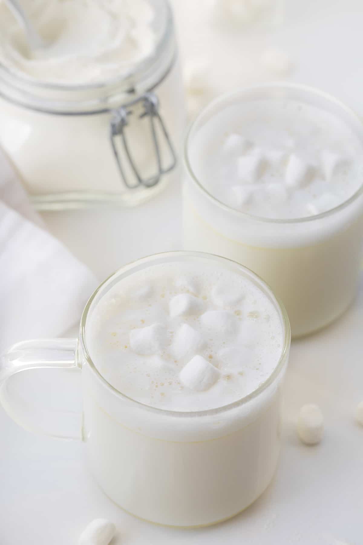 一杯白色的热巧克力放在白色的柜台上，背景是迷你棉花糖和可可。