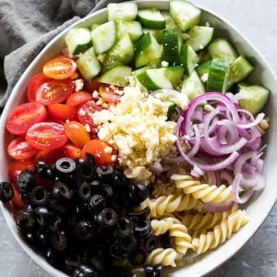 希腊意面沙拉的所有材料都放在一个白色的大碗里，边上放着灰色的餐巾gydF4y2Ba